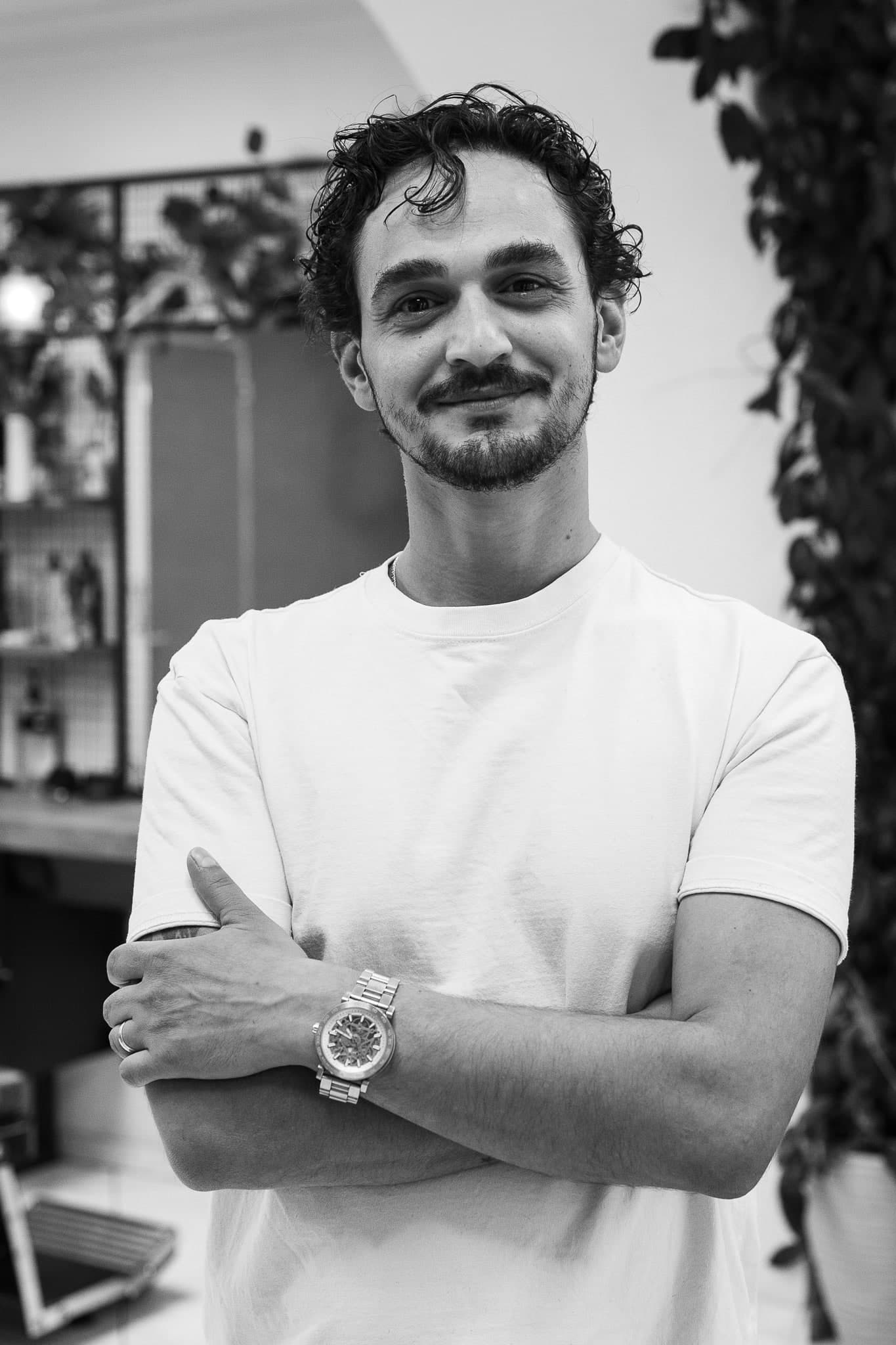 Hussein dans le salon, debout, bras croisés. Il sourit.